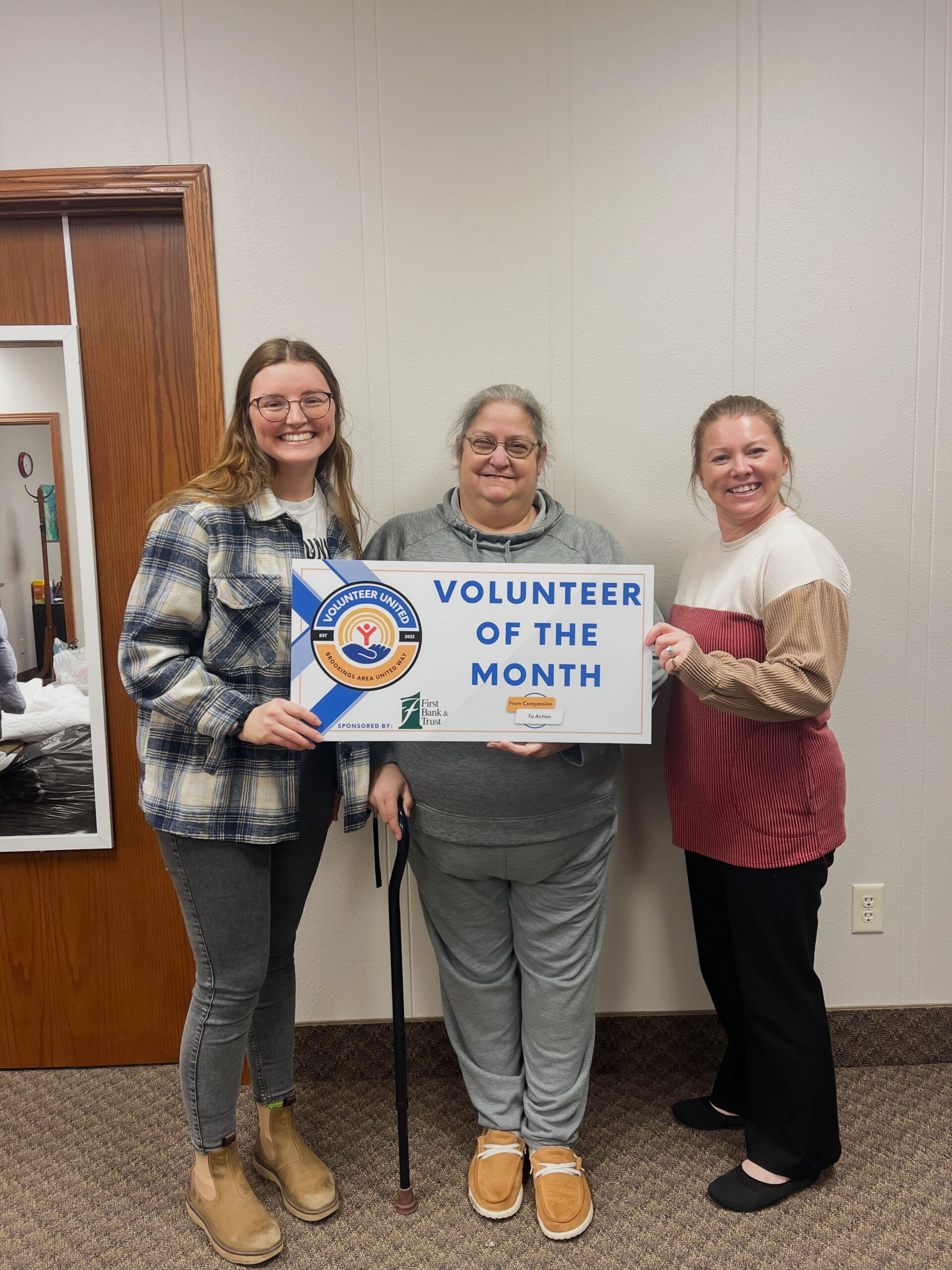 Three women smiling at the camera holding a sign that reads "Volunteer of the Month"